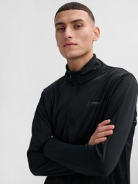 Ung mand med kort hår iført en sort sportjakke med lynlås, armene krydset, kigger selvsikkert mod kameraet mod en ensfarvet baggrund.