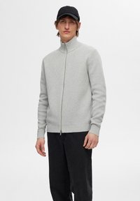 Un pull zippé gris clair en tricot texturé, avec un col montant et un ourlet côtelé, associé à un pantalon noir pour un look décontracté.