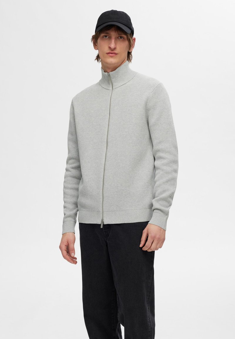Un pull zippé gris clair en tricot texturé, avec un col montant et un ourlet côtelé, associé à un pantalon noir pour un look décontracté.