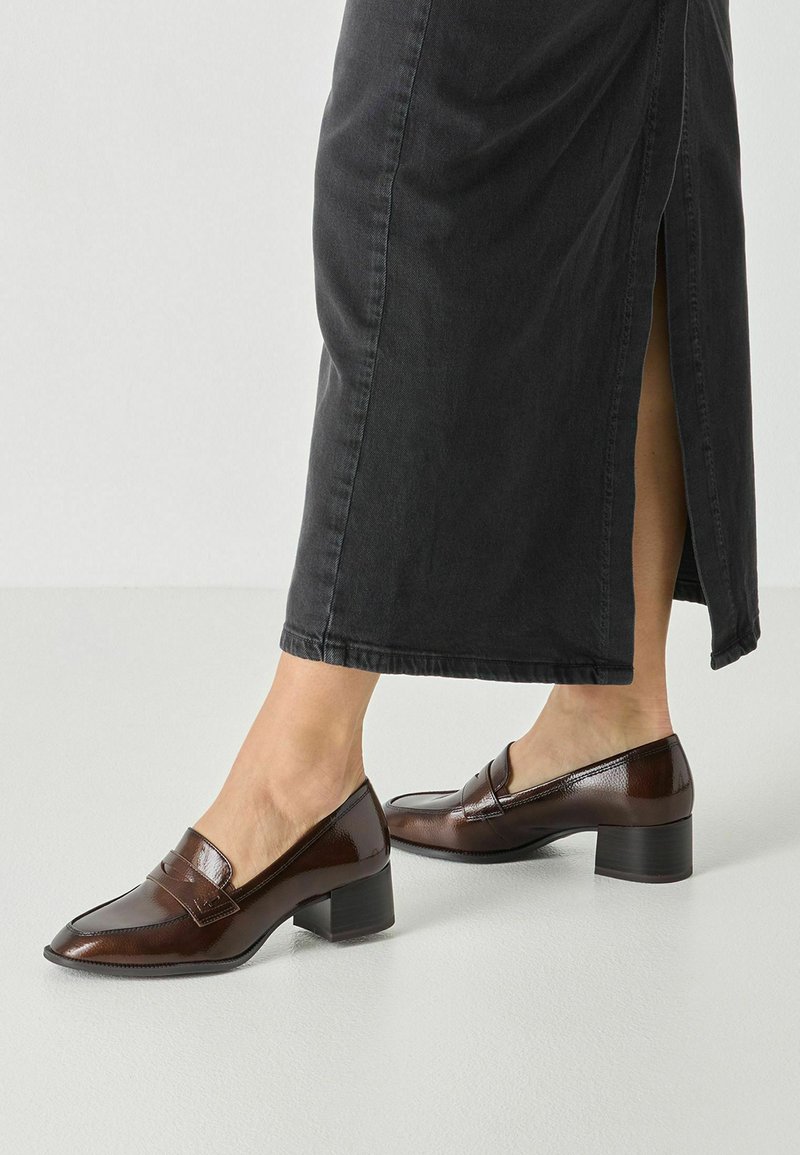 Femme portant des mocassins marron foncé à talon bas carré associés à une longue jupe noire avec une fente latérale.