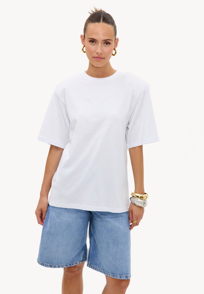 Femme portant un t-shirt blanc oversize uni et un short en jean bleu clair jusqu'aux genoux, accessoirisé avec de gros bracelets et des boucles d'oreilles créoles.
