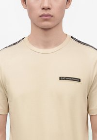 Μπεζ κοντομάνικο πουκάμισο με λογότυπα EA7 Emporio Armani στο στήθος και στις ρίγες των ώμων, φορεμένο από ένα άτομο με ουδέτερη έκφραση.