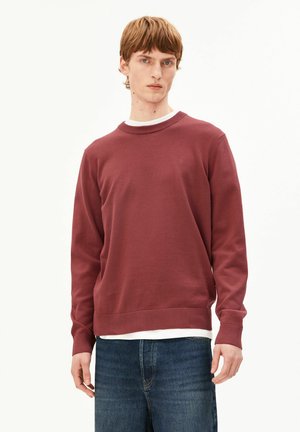 Junger Mann trägt einen kastanienbraunen Rundhalspullover über einem weißen Hemd und blauen Jeans, steht vor einem schlichten hellen Hintergrund.