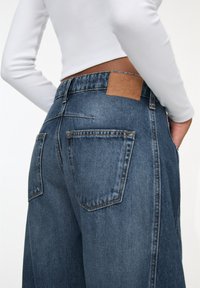Blugi din denim cu o nuanță închisă, având o etichetă din piele pe talie, buzunare standard și un design cu croială largă.
