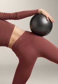 Persona attiva con abbigliamento sportivo marrone che tiene una palla da esercizio nera contro il fianco mentre si inclina di lato.