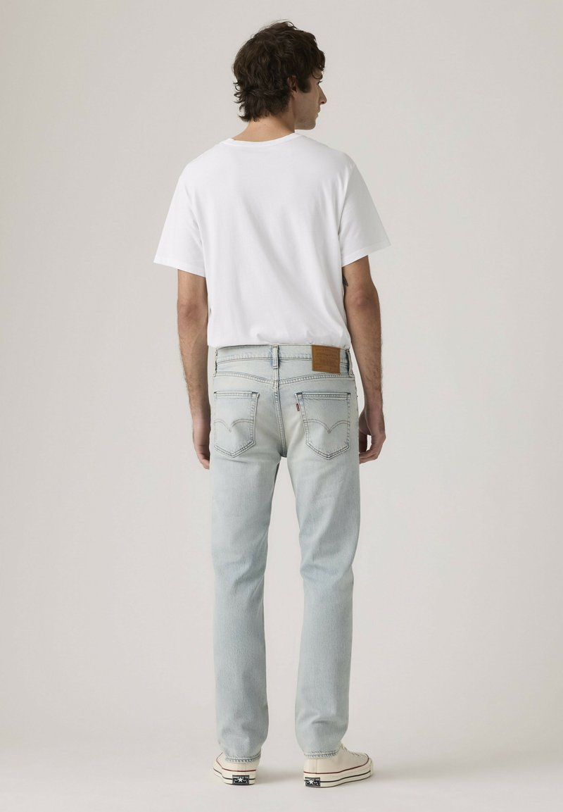 Man standing facing away wearing a plain white t-shirt, light blue jeans, and white sneakers against a plain background.