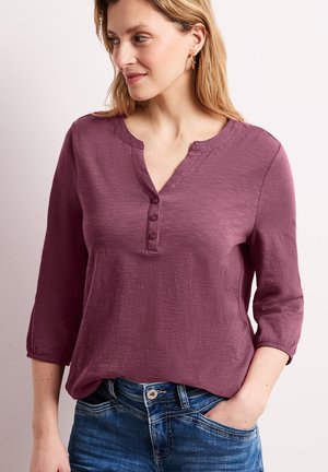 Mujer con cabello rubio que lleva una camiseta henley de manga larga color ciruela con tres botones y vaqueros azules, con la mano en el bolsillo, mirando hacia un lado.