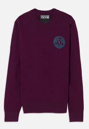 Vijoličen pleten sveter z robom na vratu in manšetes. Ima okrogel logotip na prsih z modrimi detajli. Gladka tekstura.