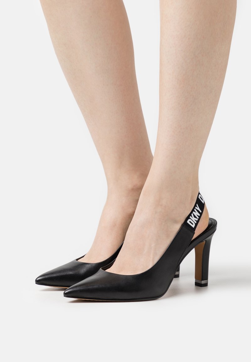 Escarpins à talons hauts noirs à bouts pointus avec brides élastiques marquées DKNY portés pieds nus sur un fond blanc.