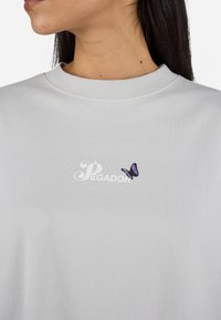 Lys grå t-skjorte med rund hals, med en hvit "Pegador" logo og en liten lilla sommerfugl grafikk på forsiden.