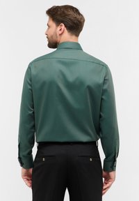 Camicia verde a maniche lunghe con una texture liscia, dotata di colletto standard e polsini con bottoni. Abbinata a pantaloni neri con doppie tasche posteriori.