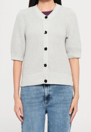 Frau trägt eine hellgraue gestrickte Strickjacke mit schwarzen blumenförmigen Knöpfen über einem dunklen Top und blauen Jeans, stehend vor einem schlichten Hintergrund.