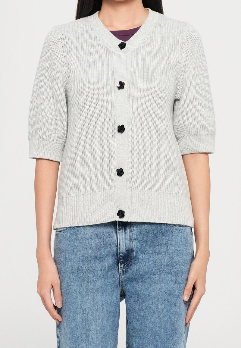 Femme portant un cardigan tricoté gris clair avec des boutons noirs en forme de fleur sur un haut sombre et un jean bleu, debout devant un fond uni.