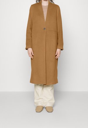 Kvinde iført en lang kamelhåret frakke med en knap, beige sweater, hvide bukser og flettede beige sko, der står mod en ensfarvet baggrund.