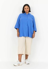 Camisa de botonadura grande en color azul con cuello, combinada con pantalones cortos crema y zapatillas ligeras. Presenta un ajuste relajado y mangas cortas.
