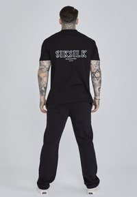 Must have lühikeste varrukatega must T-särk, millel on "SIKSILK" logo ja tekst seljal, koos sobivate mustade pükste ja valgete sneakeritega.