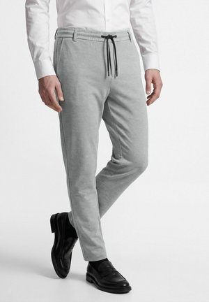 Man draagt lichtgrijze broek met trekkoord, wit overhemd, zwarte sokken en zwarte leren instapschoenen, staat met één been licht gebogen.