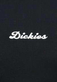 Λευκό λοφογράφο λογότυπο "Dickies" κεντημένο σε μαύρο ριμπ ύφασμα, στο κέντρο και καθαρά ορατό.