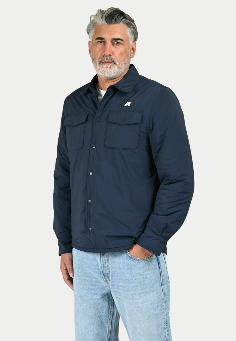 Giacca isolante blu navy con due tasche sul petto, chiusura con bottoni e una logo accentuato sul lato sinistro. Indossata con jeans chiari.