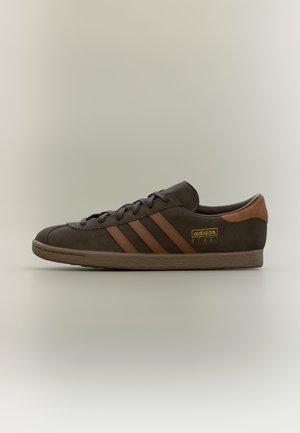 Baskets Adidas Stadil marron avec trois bandes, lacets et un patch talon marron clair sur un fond clair uni.