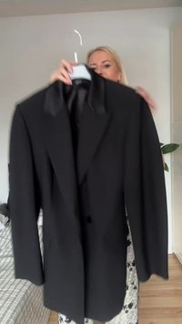 Black blazer with notched lapels, single button closure, long sleeves, and smooth texture. Hangs on a hanger against a light background.