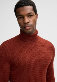 Maglione a collo alto di colore arancione bruciato con un motivo strutturato e colletto a costine. Il design aderente mette in risalto la forma e i dettagli del tessuto.