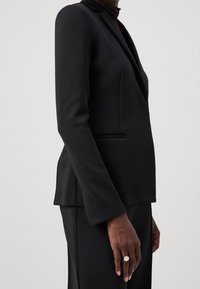 Svart blazer med en strukturerad form, en knapp stängning och sidofickor. Slät tyg och skr äddarsydd passform, tillsammans med smala svarta byxor.