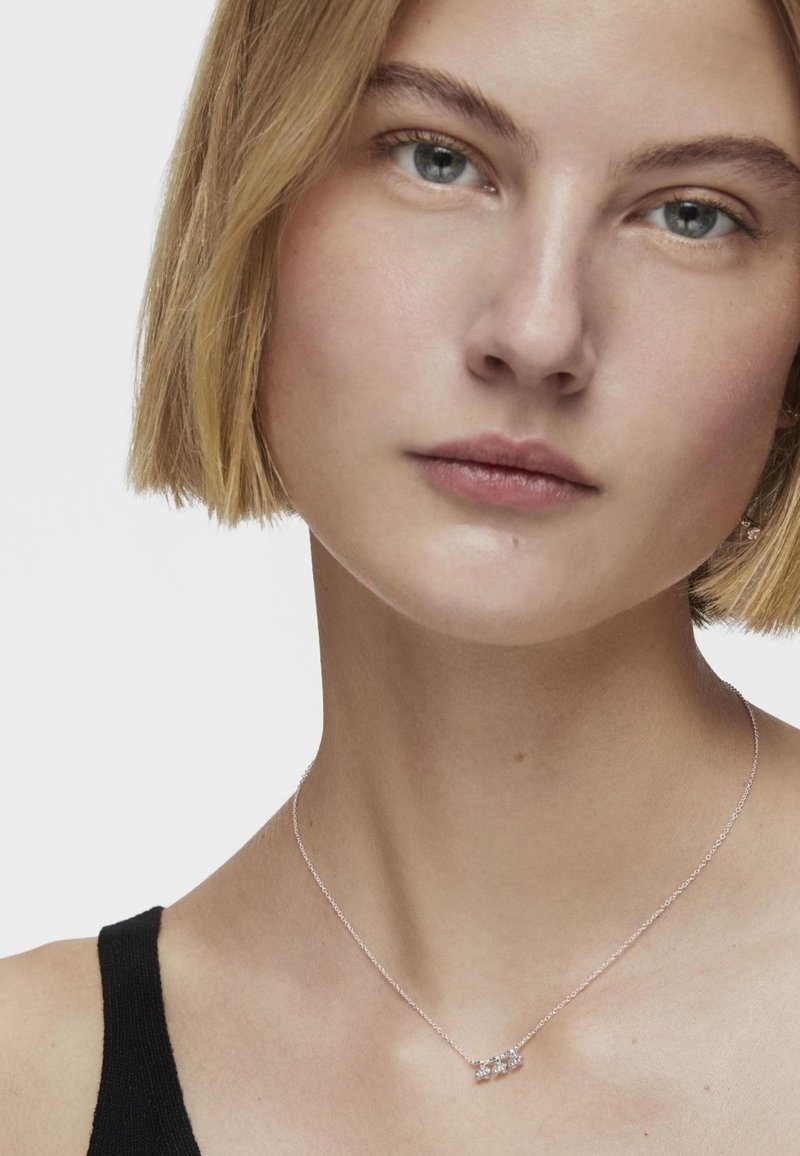 Collar de plata con tres pequeñas piedras transparentes, llevado en un cuello desnudo, sobre un fondo liso, combinado con una blusa negra sin mangas.