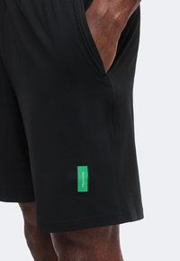 Gros plan d'une personne portant un short noir avec une étiquette rectangulaire verte sur la jambe gauche et la main dans la poche latérale.