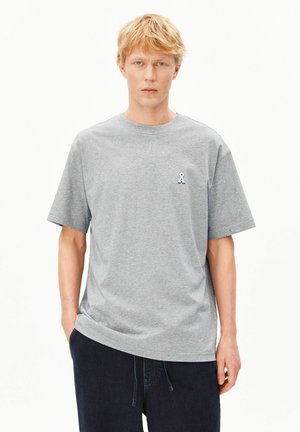 Uomo biondo in piedi con espressione neutra, indossa una maglietta grigia semplice a maniche corte con un piccolo logo a nastro blu sul petto e pantaloni scuri.