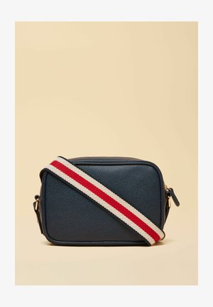 Obdĺžniková tmavomodrá kožená kabelka na crossbody s širokým látkovým pásom v pruhovanom červenom a bielom prevedení na béžovom pozadí.