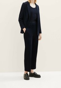 Donkerblauwe blazer over een bijpassend tanktop, gecombineerd met rechte broekspijpen en zwarte instapschoenen. Glad materiaal met een getailleerde pasvorm.