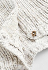 Beige gestrickter Pullover mit einem strukturierten Design, ausgestattet mit einem Holzknopf und einem Etikett. Der Stoff hat ein weiches, kräftiges Aussehen.
