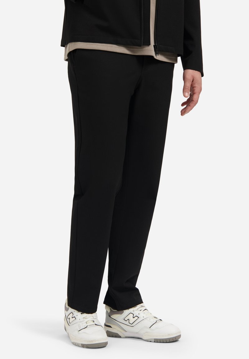 Zwarte, taps toelopende broek met een soepele textuur en zijnaad, gecombineerd met witte sneakers met grijze accenten en veters.