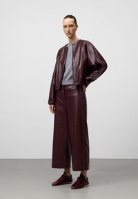 Giacca bomber in pelle bordeaux con orlo a costine, abbinata a pantaloni a gamba larga coordinati. Maglione grigio aderente sotto e sneakers bordeaux.