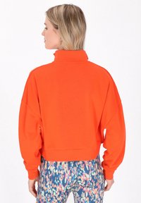 Rövidített narancsszínű pulóver, magas gallérral és laza ujjal, színes mintás leggingsel párosítva. Simas látványú anyag.