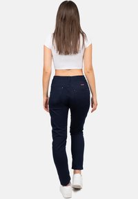 Femme aux longs cheveux bruns portant un crop top blanc, un pantalon bleu marine et des baskets blanches, se tenant de dos.