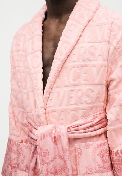 Άτομο με σκούρο δέρμα που φοράει ρόμπα ροζ με υφή και ανάγλυφο κείμενο "Versace" και μια μοτίβο ζώνη από μετάξι δεμένη στη μέση.