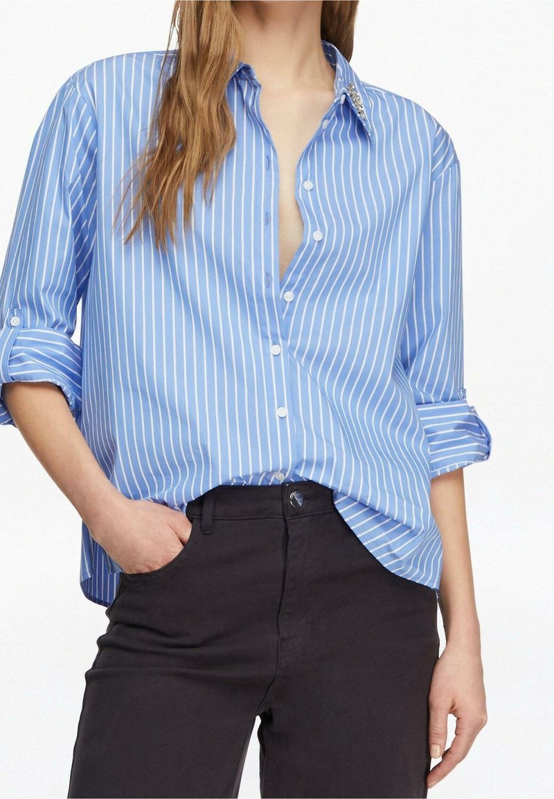 Femme portant une chemise boutonnée à rayures verticales bleues et blanches avec les manches roulées et un pantalon noir, une main dans la poche, debout devant un fond blanc.