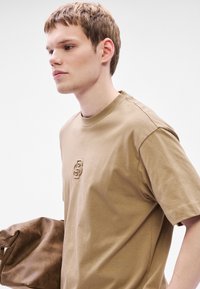 Μπεζ βαμβακερό T-shirt με στρογγυλή λαιμόκοψη και κοντά μανίκια, με ανάγλυφο λογότυπο στο στήθος. Το μοντέλο κρατάει καφέ τσάντα.