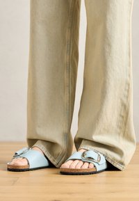 Sandálias azul claro feitas de material suave, com uma tira larga e um grande fecho de metal prateado, assentadas numa palmilha de cortiça com sola texturizada.