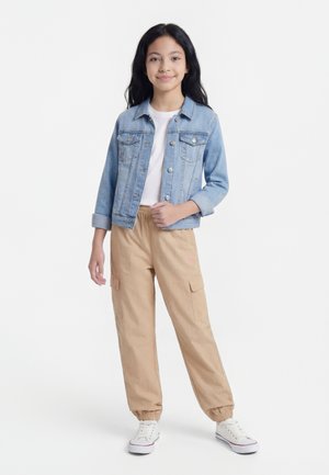 Κορίτσι με μακριά σκούρα μαλλιά, φορά ανοιχτό μπλε τζιν μπουφάν, λευκό πουκάμισο, μπεζ παντελόνια cargo και λευκά αθλητικά παπούτσια, στέκεται με το ένα χέρι στο μπουφάν.