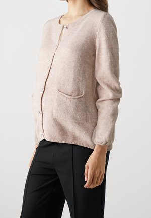 Femme portant un cardigan boutonné beige clair avec de petites poches et un pantalon noir, se tenant contre un fond blanc uni.