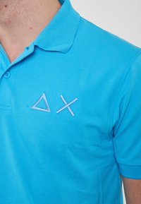 Polo shirt azzurro chiaro con un tessuto testurizzato; presenta un triangolo e una croce ricamati in una tonalità leggermente più chiara.