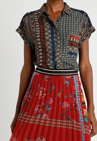 Chemise à manches courtes à motifs avec des accents bleus et une poche plaquée, associée à une jupe plissée rouge ornée de broderies florales et colorées.