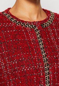 Rode textuurjacket met zwarte en gouden kettingrand langs de halslijn en aan de voorkant, gedragen door een persoon die alleen de nek en bovenste borst laat zien.