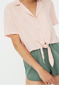 Short-sleeve, tie-front shirt in light pink fabric with a collar and white buttons, paired with green shorts. Soft texture, relaxed fit.