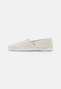 Sapatos slip-on em lona bege com uma textura suave, painéis laterais elásticos e solas de borracha branca, apresentando um design minimalista e bico arredondado.