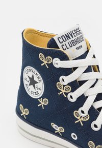 Baskets montantes en toile bleu marine avec des lacets blancs, des accents jaunes et un motif en diamant. Présente des designs de raquettes de tennis et un patch avec le logo.