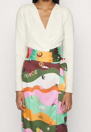 Frau trägt ein weißes Langarm-Wickeltop und einen bunten Wickelrock mit Aufdrucken von Frieden, Liebe, Sternen, Vögeln und abstrakten Formen.